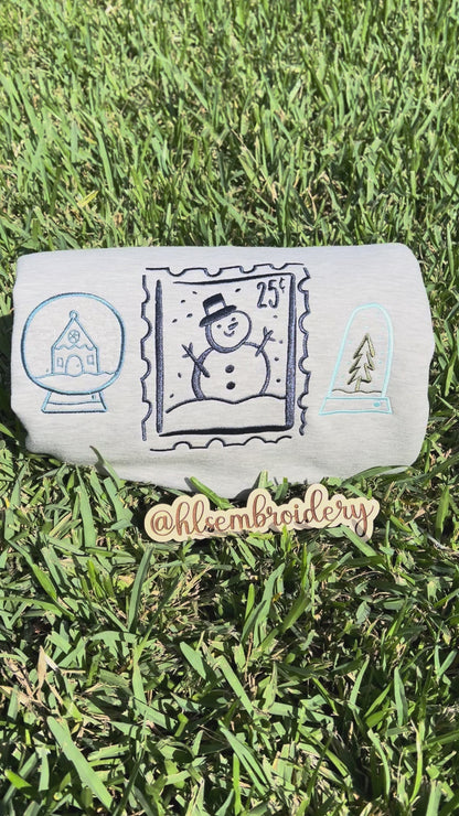 Embroidered designs of a snowman, snow globe, and pine tree on grass.