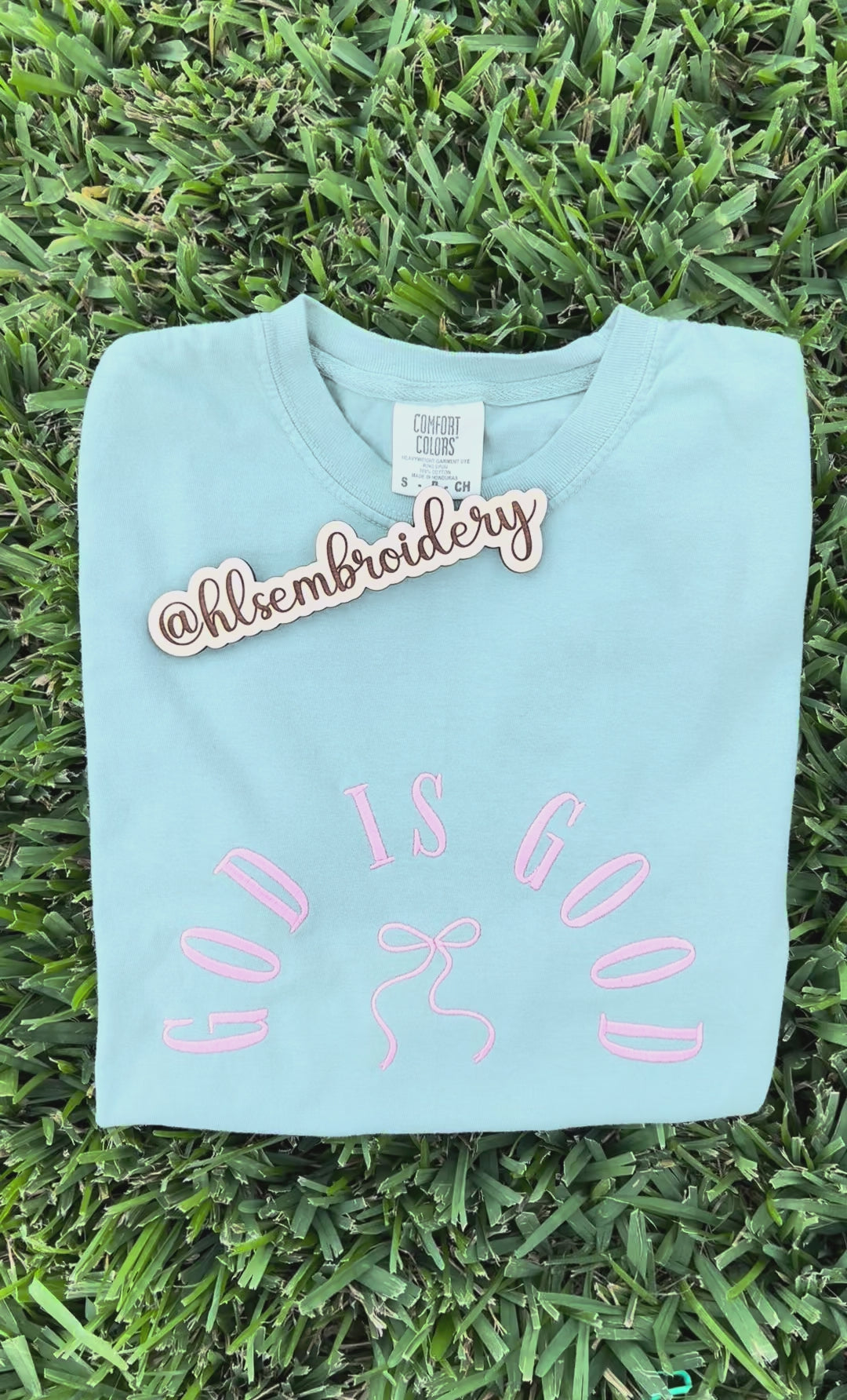 Light blue t-shirt with 'GOD IS GOOD' text and bow design on grass