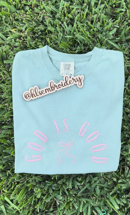 Light blue t-shirt with 'GOD IS GOOD' text and bow design on grass